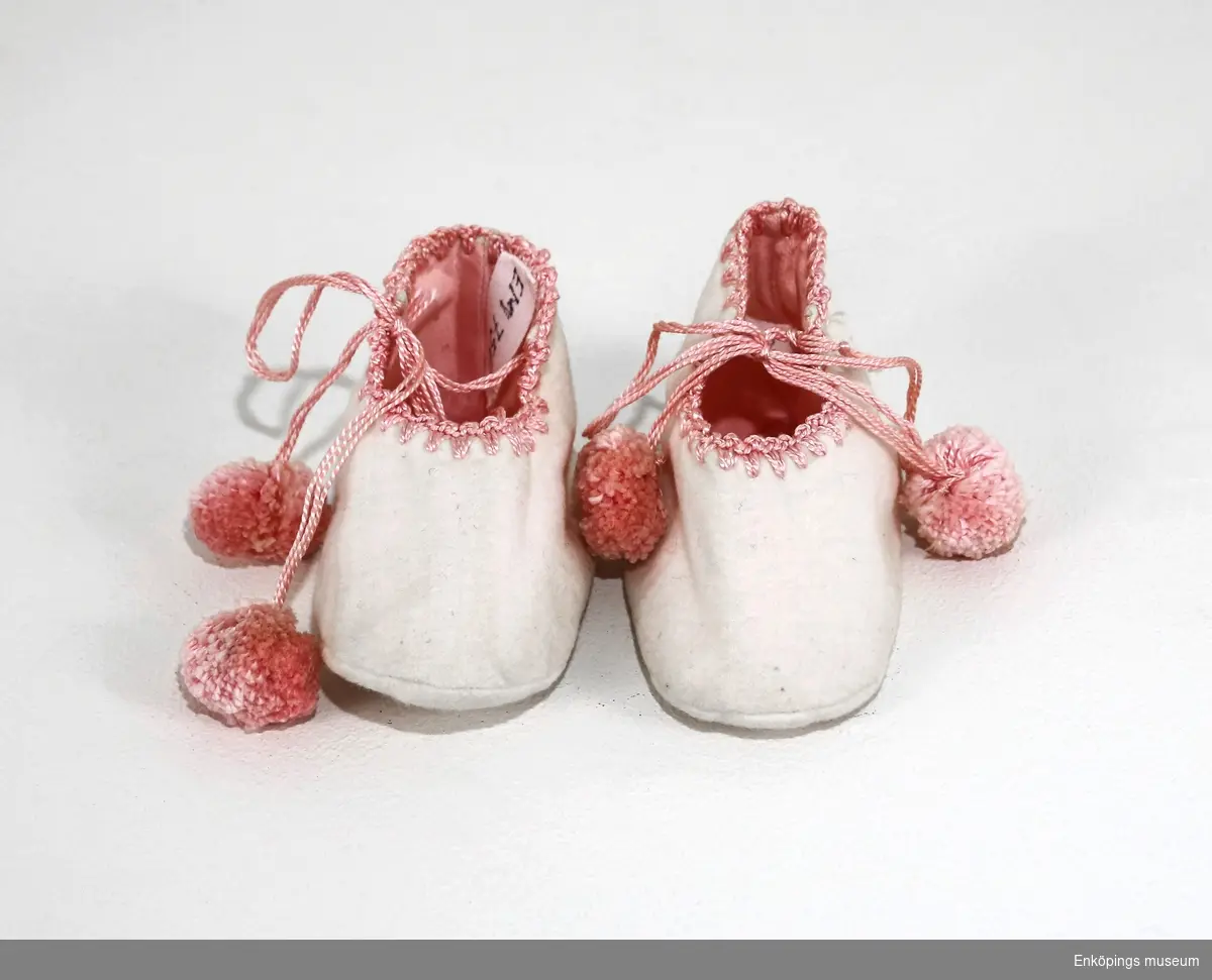 Vita flanellskor med rosa foder, toffelmodell. Skorna har virkad kant i rosa och knytband med bolltofsar. Tofflorna är tillverkade 1922.