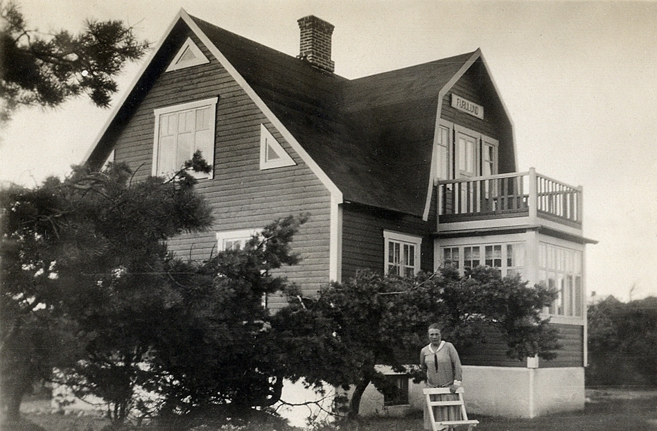 Ett bostadshus med glasveranda och balkong. Ovanför balkongdörren syns en skylt med texten: "Furulund".
Framför huset står en okänd kvinna bakom en trädgårdsstol.