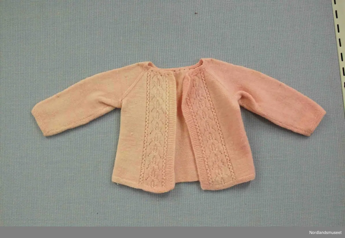 Lys rosa strikkejakke til spedbarn. Flettestrikk i to rader foran. Åpning foran. Fargen er noe falmet.