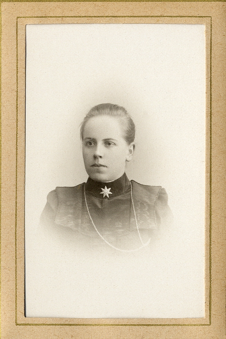 En okänd ung kvinna, klädd i tvådelad klänning med hög sammetskrage. Vid kragens syns en brosch och en smal klockkedja.
Bröstbild, halvprofil. Ateljéfoto.