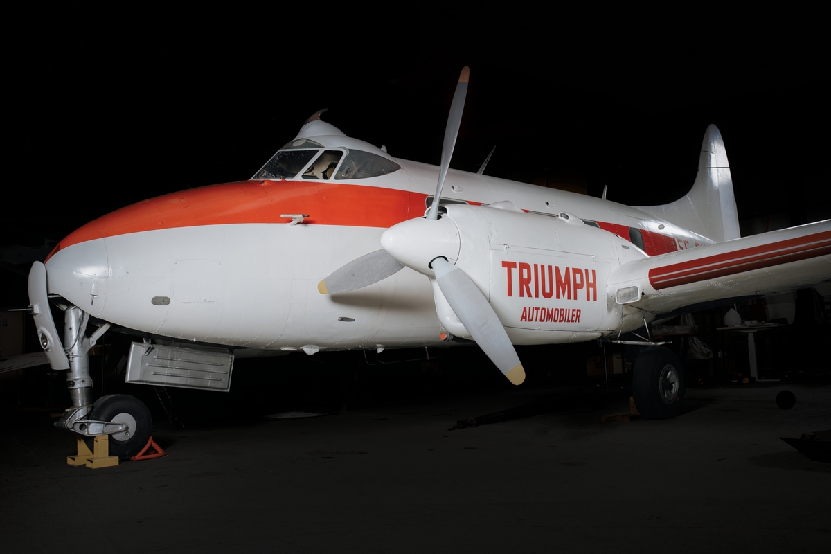 Flygplan av modell DH 104 Dove. Tvåmotorigt, lågvingat propellerflygplan av lättmetall. Vitmålat med röd registrering och dekor. Påskrift ”Triumph”.