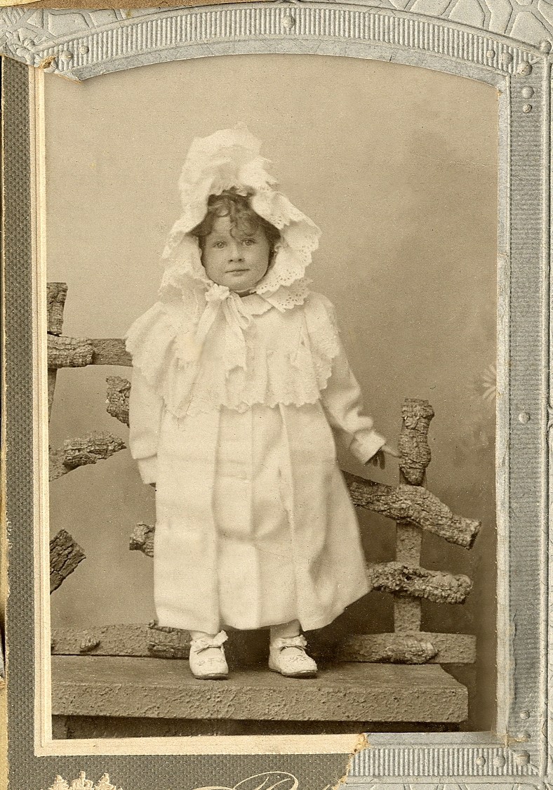 En liten flicka i lång vit kappa och en stor hätta med brodyr. Hon står på en bänk framför en "gärdesgård". 
Helfigur. Ateljéfoto.

Fotografens dotter.