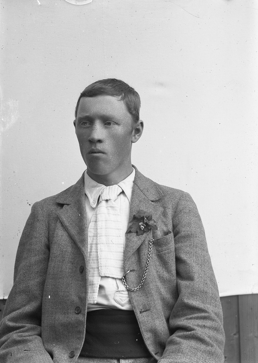 Ung mann med lys jakke, klokkekjede, blomst på jakkeslaget og bredt magebelte med monogram(?).