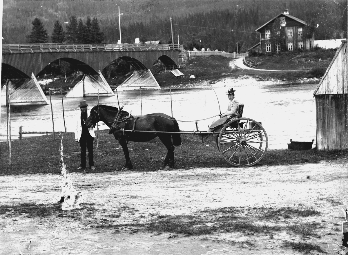 Nybergsund, bildet er tatt på fotografens egen gårdsplass. Kvinne sitter i en gigg, hest, kjerre, bru, elv.