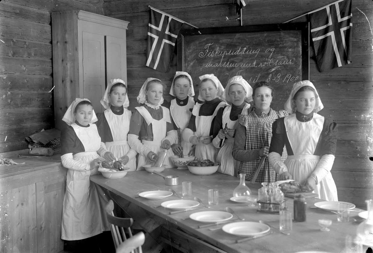 Åtte jenter på husstellkurs foran tavle der det står "Fiskepudding og marssne med rød saus. Trysil 3 -3 1900".