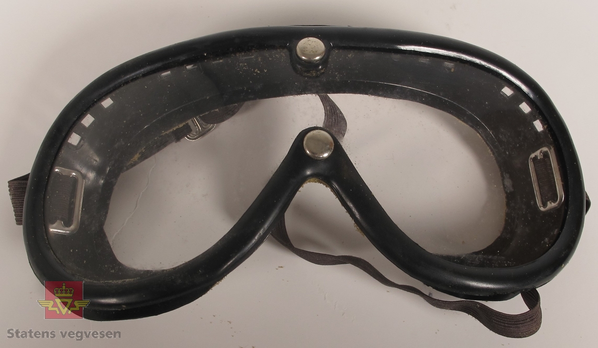 Briller i svart gummimateriale, med plastvindu som er festet med til brillen med to nagler. Det er rester etter skumgummi på den siden som er mot ansiktet. I gummien på siden og på toppen rundt brillen er det luftehull. Justerbart strikk festet til hver siden av brillen for justering til hode eller hjelm.