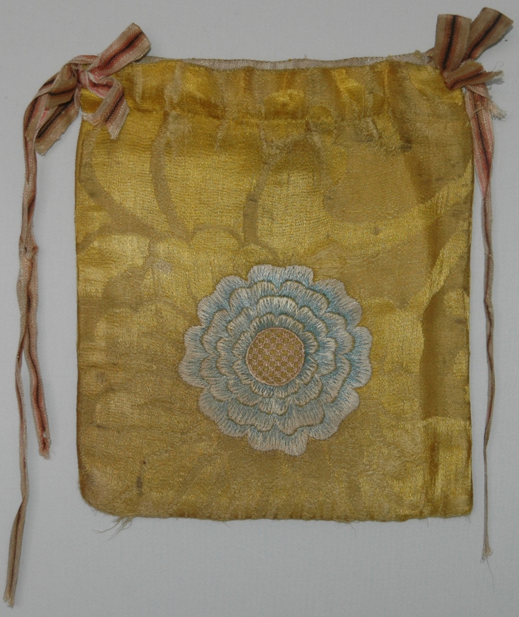 Handväska av gult sidendamasttyg med broderi i flera färer. Märkt på ena sidan BCB 1787 i ett hjärta under en grevlig krona. På andra sidan en broderad blomma. Fodrad med linnetyg.