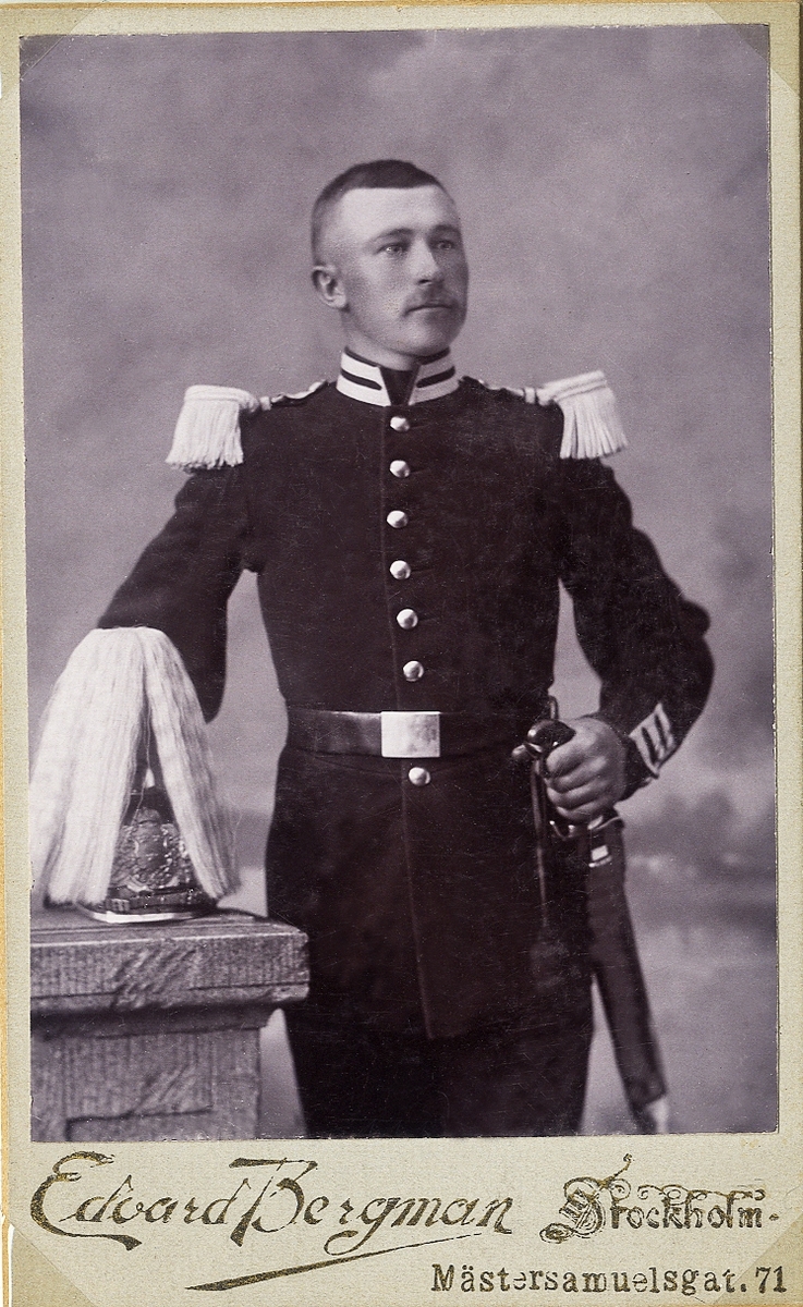 Porträttfoto av en man i militär paraduniform. På pelaren bredvid ligger en kask med tagelplym. 
Knäbild, halvprofil. Ateljéfoto.