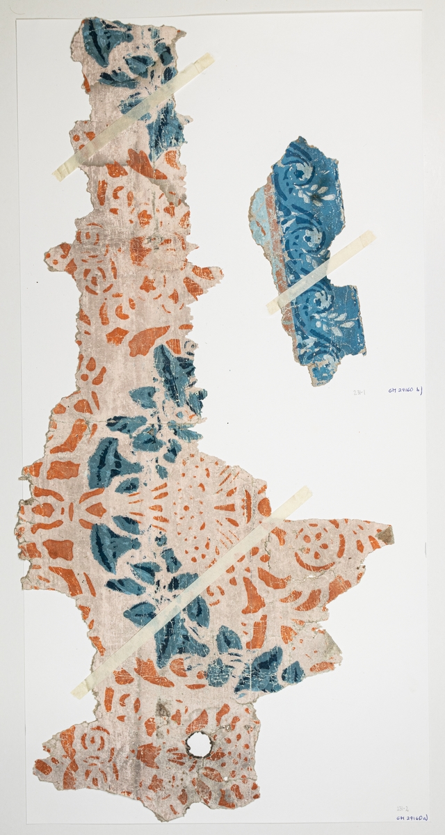 Fragment av schablonmålad tapet med en girland av klockformade blommor i två nyanser av blått kring en stiliserad urna i orange på ljust rosa botten.