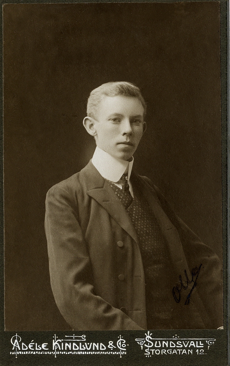 Porträttfoto av en ung man i mörk kavajkostym med prickig väst och stärkkrage med fluga. 
I nedre högra syns (otydligt) autograf: "Olle".
Midjebild, halvprofil. Ateljéfoto.