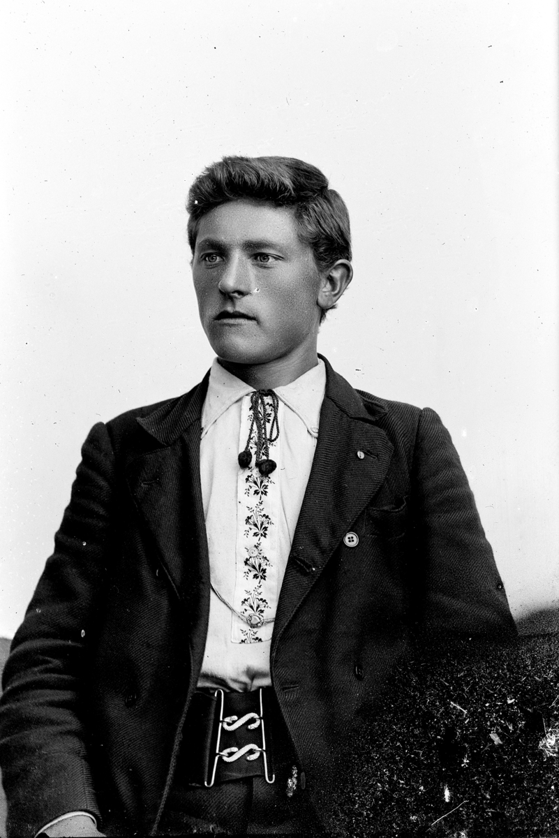 Studioportrett, ung mann, med brodert skjorte, "lisse", lissebinding i halsen, mørk jakke, bredt belte med to S'hekter. Svensk?