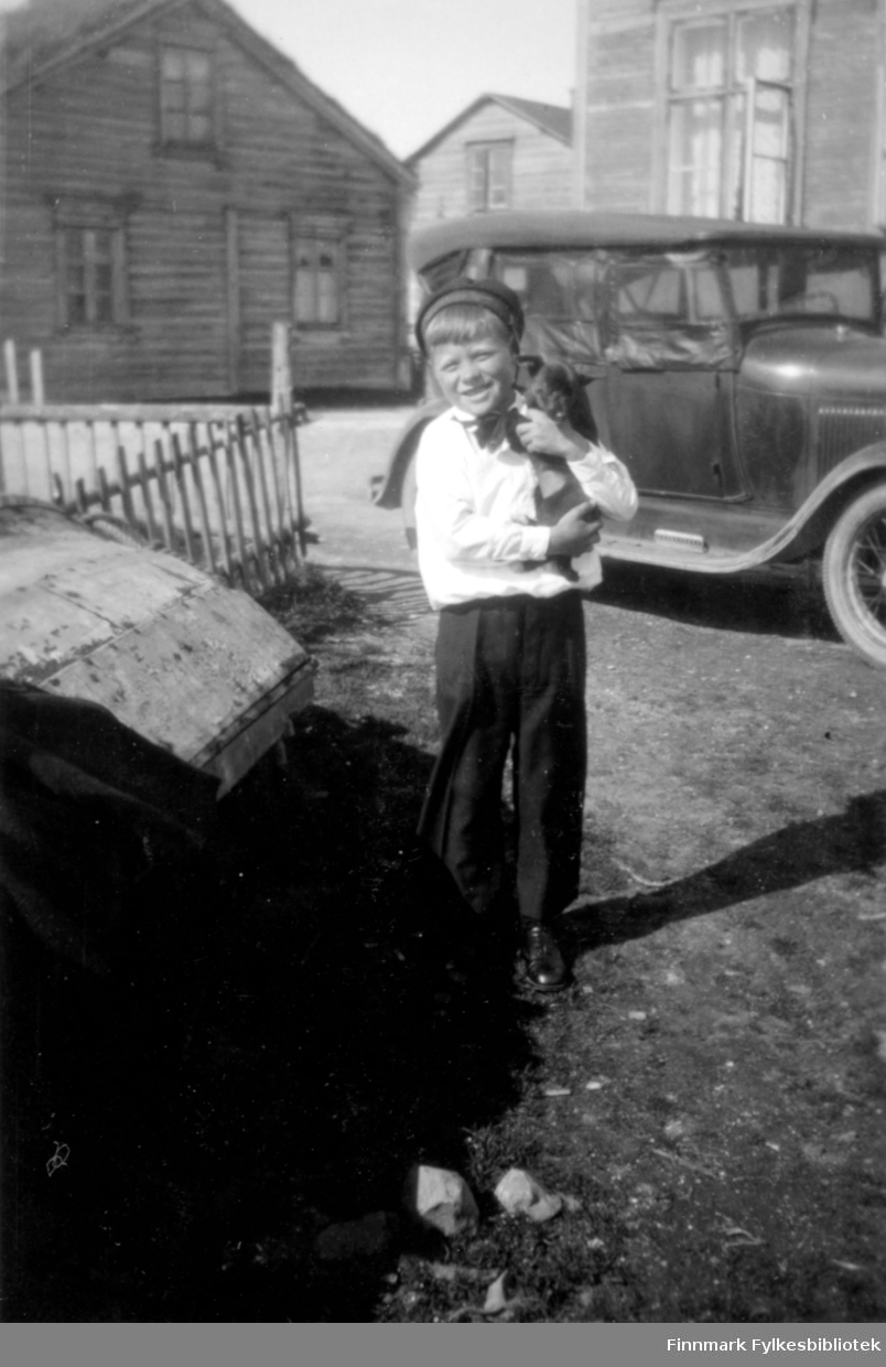 En gutt holder i hånden en hundevalp. Stedet er antakelig Salttjern. En bil parkert foran et hus.