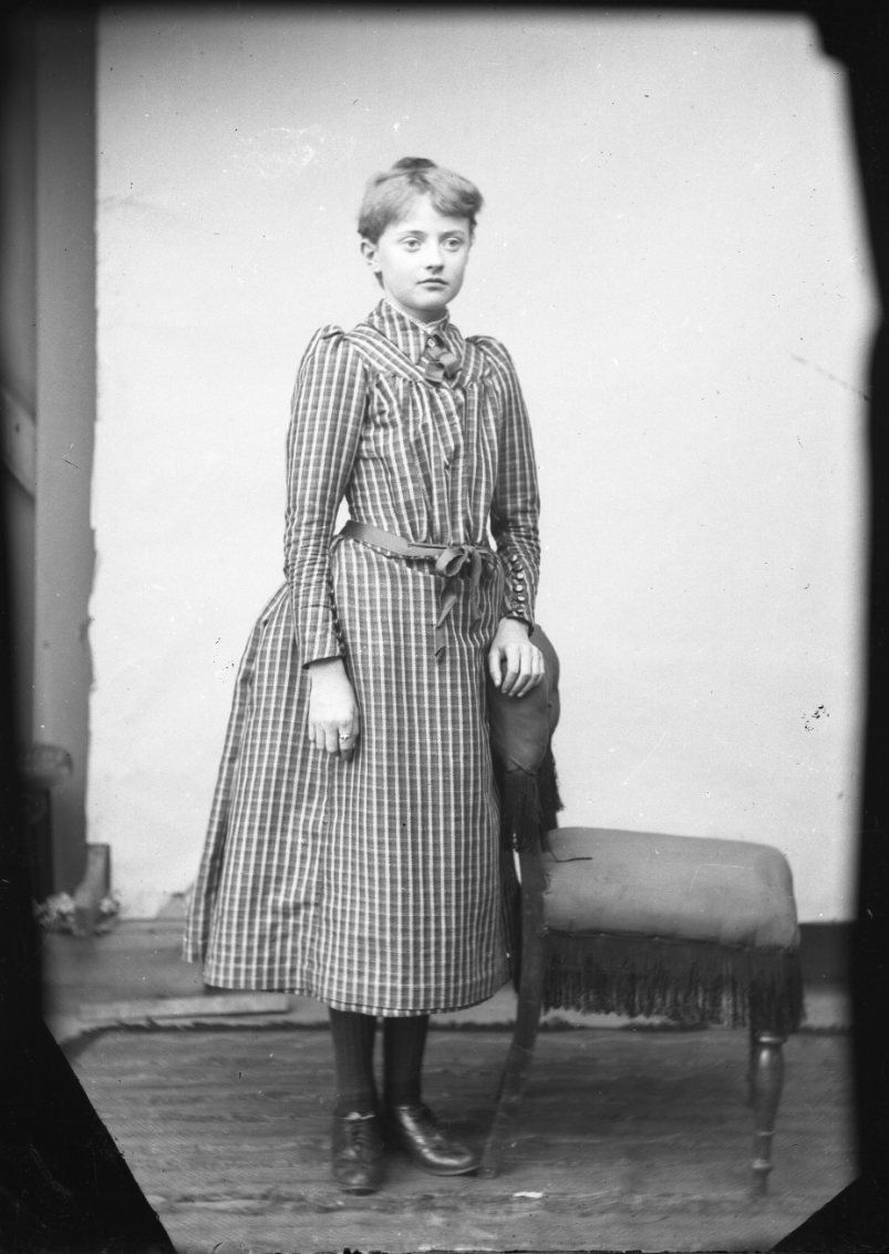 Porträtt av Ester Grönberg. Hon står med vänster hand vilande på en stolsrygg. Hon bär en rutig klänning.