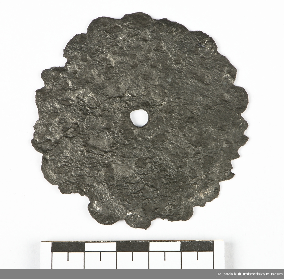 En cirkelformad platta med 18 stycken runda taggar i kanten. I metallen syns rester av mönster och i mitten finns ett hål. Troligtvis fårn likkista.