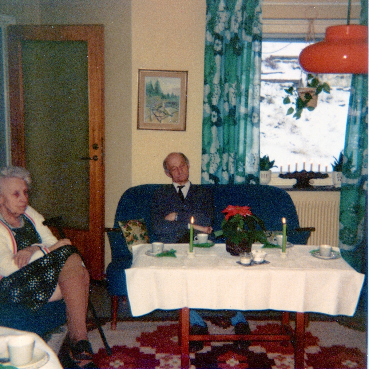 Två äldre personer sitter runt ett bord i Brattåshemmets dagrum på nedre våningen julen 1979. Okänd kvinna till vänster. Mannen i mitten är Anselm Zackrisson (1900 - 1987). Genom fönstret ses snö utanför.
