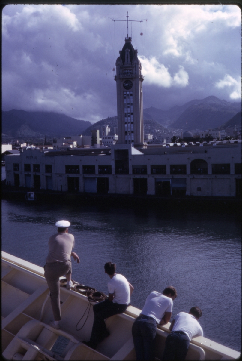 Mannskap i baugen på cruiseskipet 'M/S Sagafjord' ved innseiling til Hawaii, med Aloha Tower i bakgrunnen. 'Sagafjord' Around The World via Africa Cruise 1966.