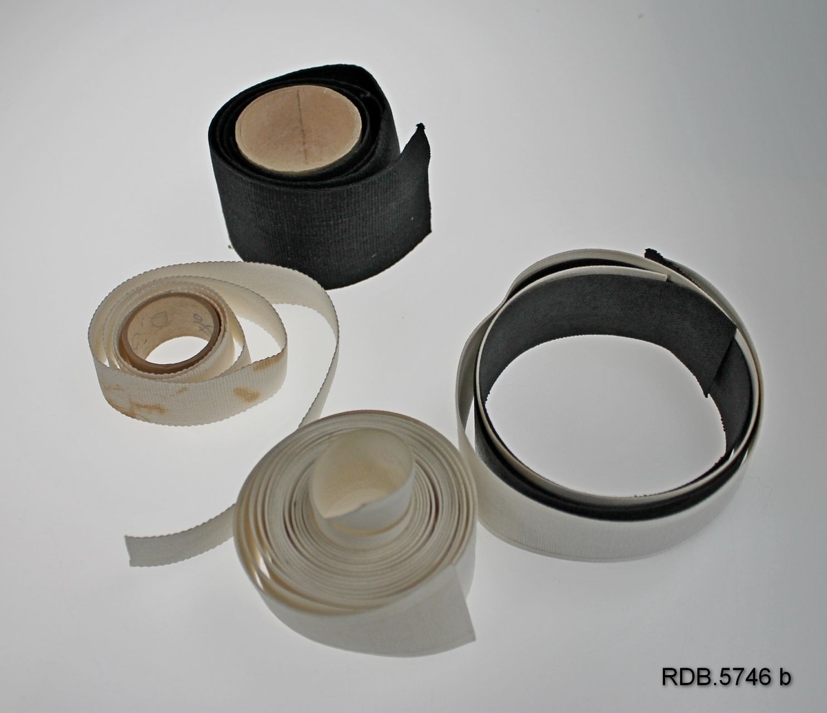 En liten pappeske eske uten lokk som inneholder 3 ruller og 2 biter stivt innleggsband. 2 kvite og 1svart rull, 1 svart og 1 kvit bit.