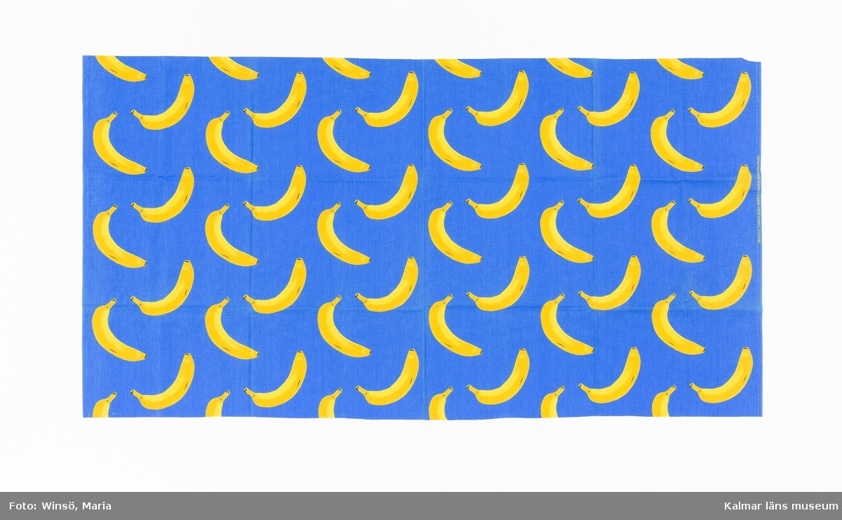 KLM 46157:690. Tyg. Metervara. Tryckt mönster på vit tuskaftad bomull. Mönster med gula bananer mot blå botten. Tryckt gul text i höger stadkant: "BANANA" CARINA HÄGG DESIGN. Printed by Rydboholms Sweden.
