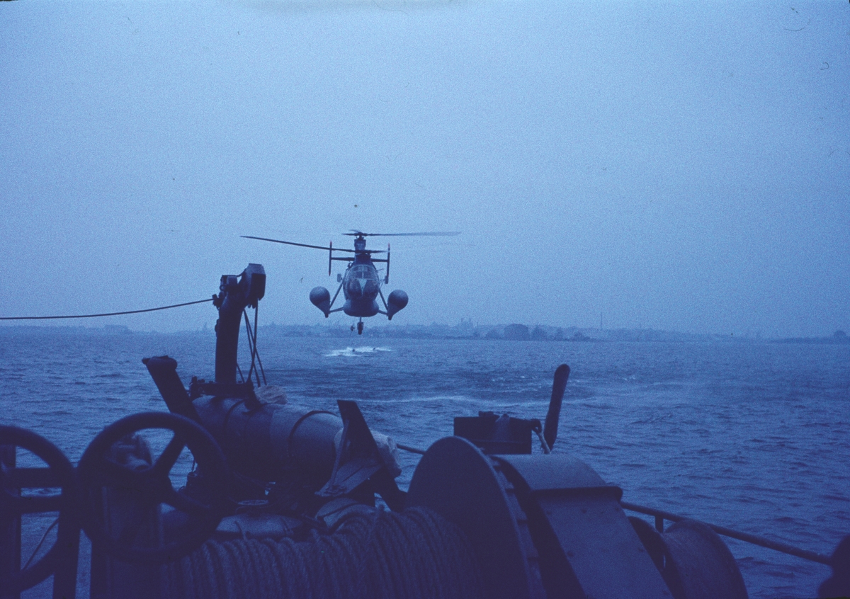 Bilden föreställer en så kallad flygande banan - en helikopter typ Vertol 44 under flygning. I förgrunden ser man akterdäcket av en minsvepare. I bakgrunden anar man siluetten av Karlskrona.