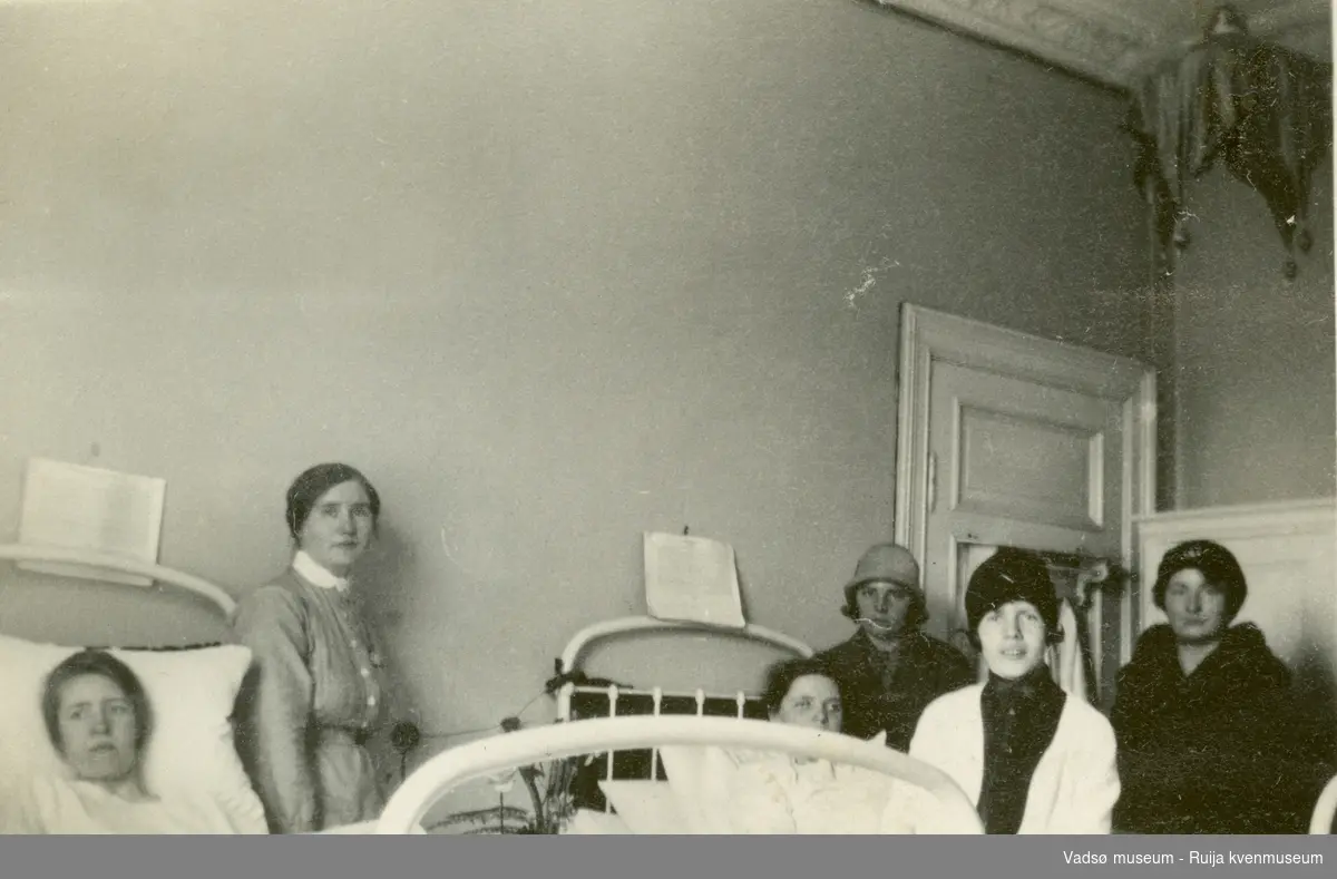 Gruppe unge kvinner samlet ved sykesenger. Mathilde Smedstad ligger i sengen til høyre. Antatt Vadsø sykehus 1924.