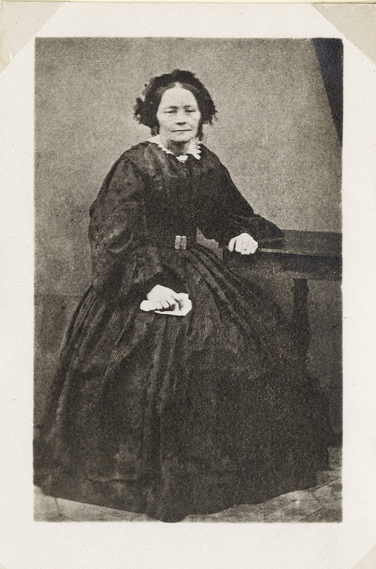 Porträttfoto av kvinna i mörk krinolin med mörk hårklädsel på huvudet. Hon sitter vid ett pelarbord som hon lutar ena armen mot. 
Helfigur, en face. Ateljéfoto.