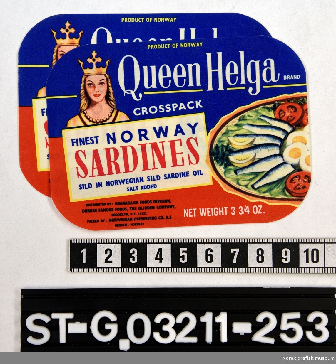 Etiketter i rødt og blått, med en fremstilling av en kvinne med krone på venstre side av etiketten, og et fat med fisk, egg og grønnsaker på høyre side. 

"Finest Norway sardines in Norwegian sild sardine oil"