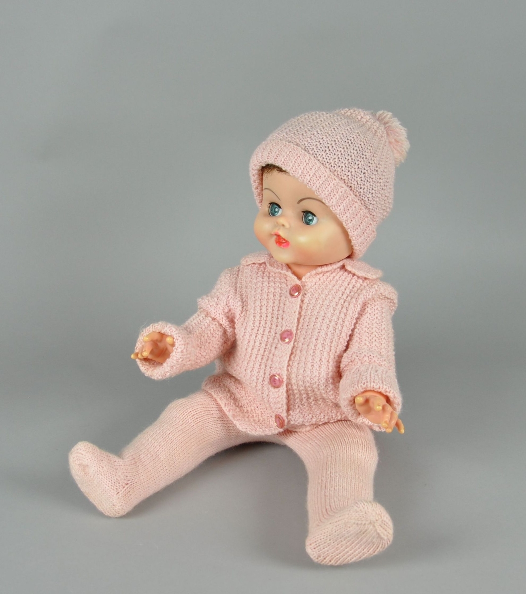 Dukke av plast. Dukken har bevegelige øyelokk og lemmer. Dukken er kledd med bukse, genser og lue av strikket, rosa tekstil. På genseren er det fire blake knapper, luen har en dusk.
