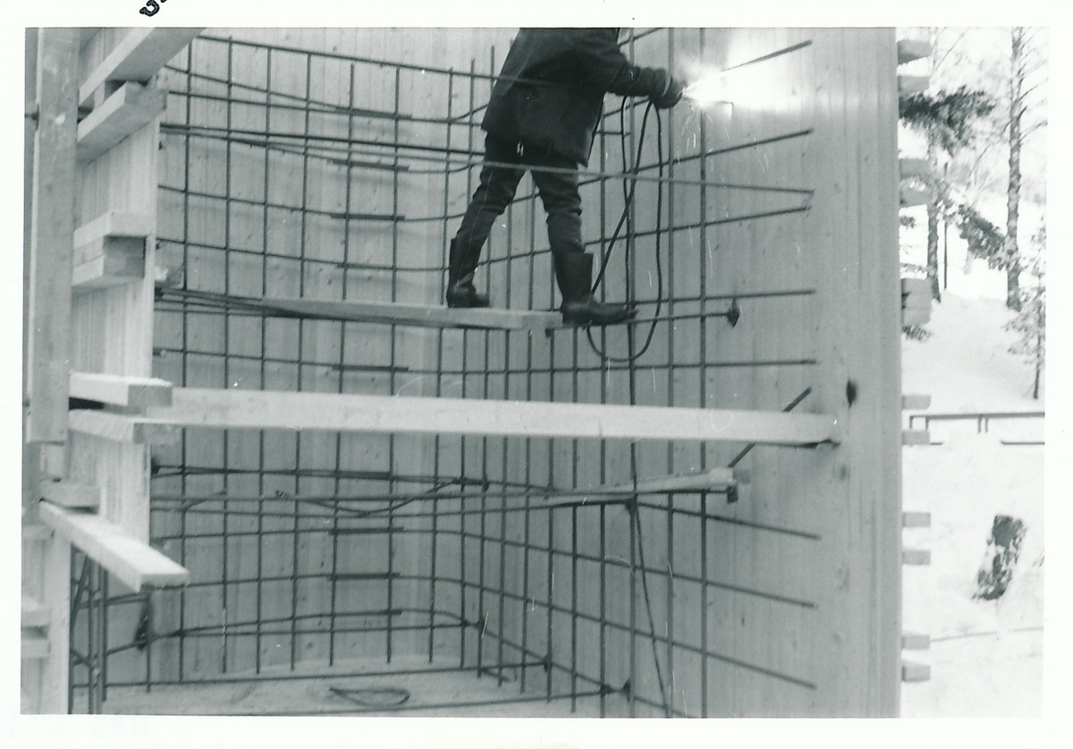 Mann gjør sveisearbeid på armeringsjern inne i trekonstruksjon som har tre vegger.