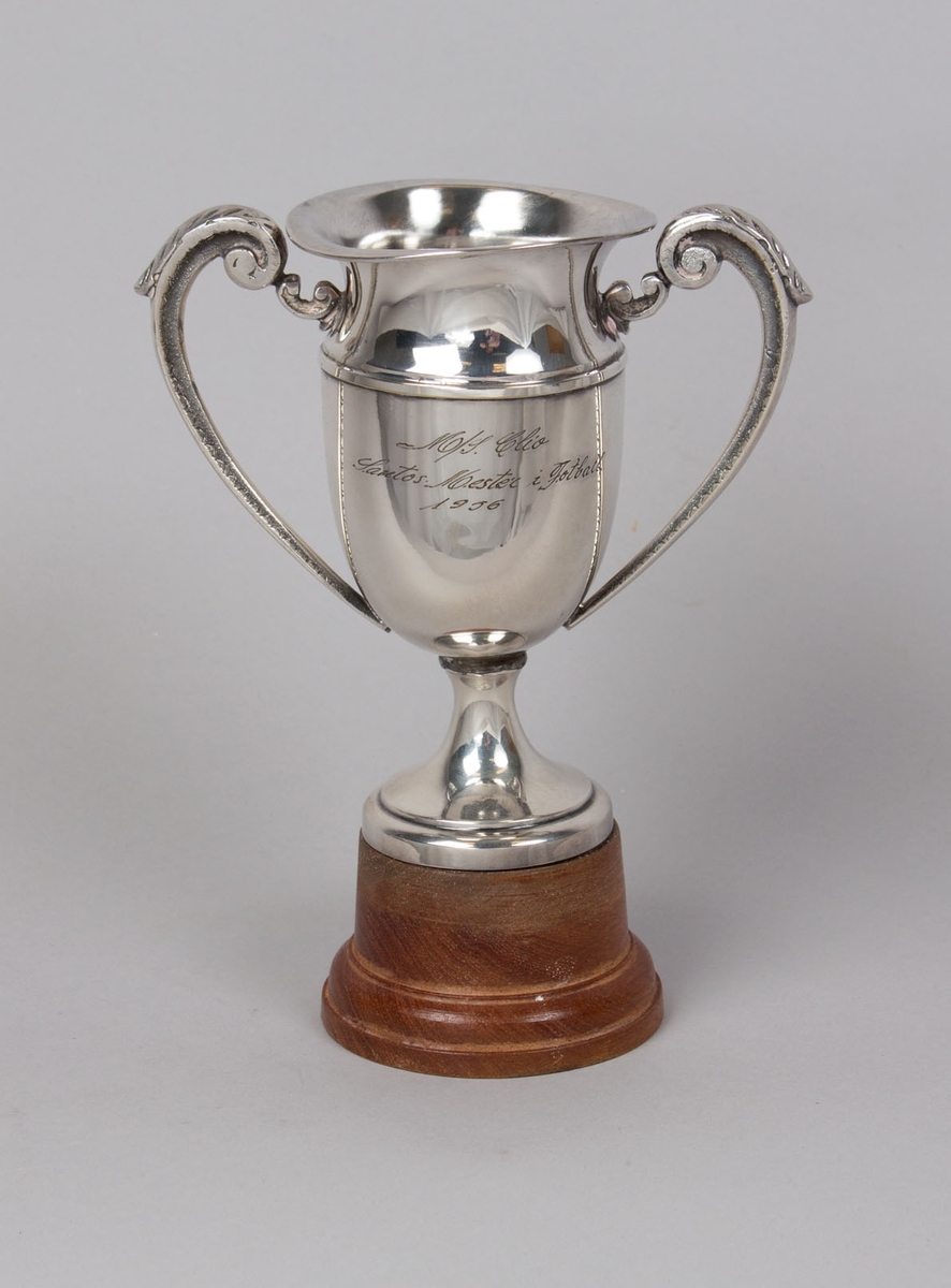 Sølvpokal tildelt MS CLIO for mester i fotball 1956, konsul Olav Syrdahls ærespris. Pokal med hank på hver side montert på trestett. 
