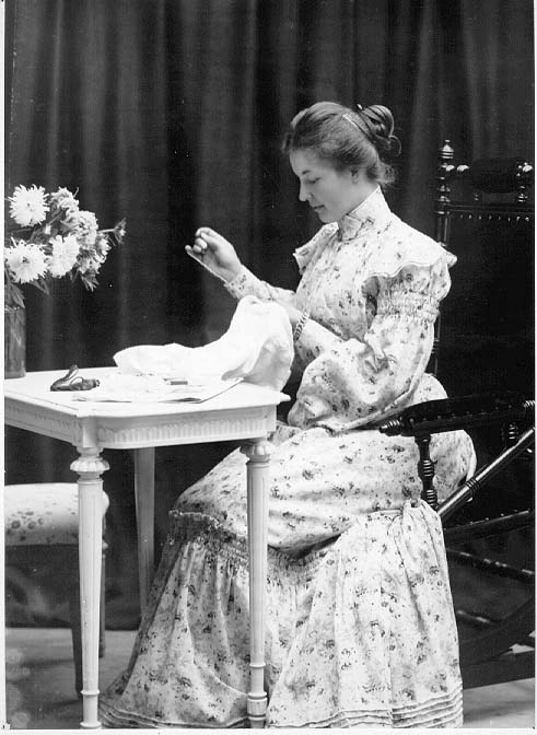 Fru Malmqvist i lång blommönstrad klänning sitter vid ett bord och broderar. På bordet framför henne ligger det en tidning. Där står också en vas med blommor.