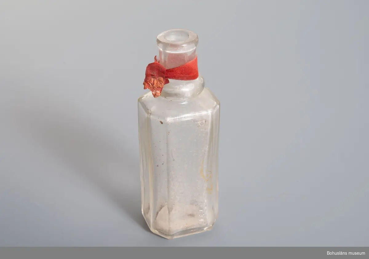 Flaskan rektangulär med två fasade kanter, en sida halvrund. 
Halsen har en nod samt rött band.