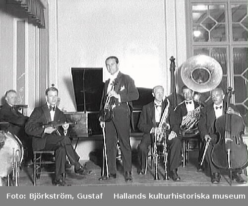 Okänd orkester med sex män som spelar på Societeten i Varberg. De poserar med flygel, fiol, saxofon, bastuba och cello men även trumma, trumpet, kontrabas och klarinetter finns med i bild.