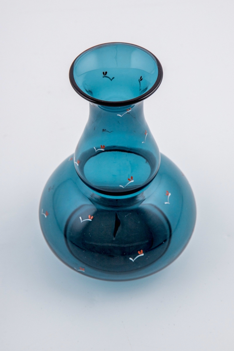 Kalebassformet vase i gjennomskinnelig blålig glass. Korpus hviler på en sirkulær fot. Vasen er dekorert med emaljemaling, iform av stiliserte grener med bladverk.