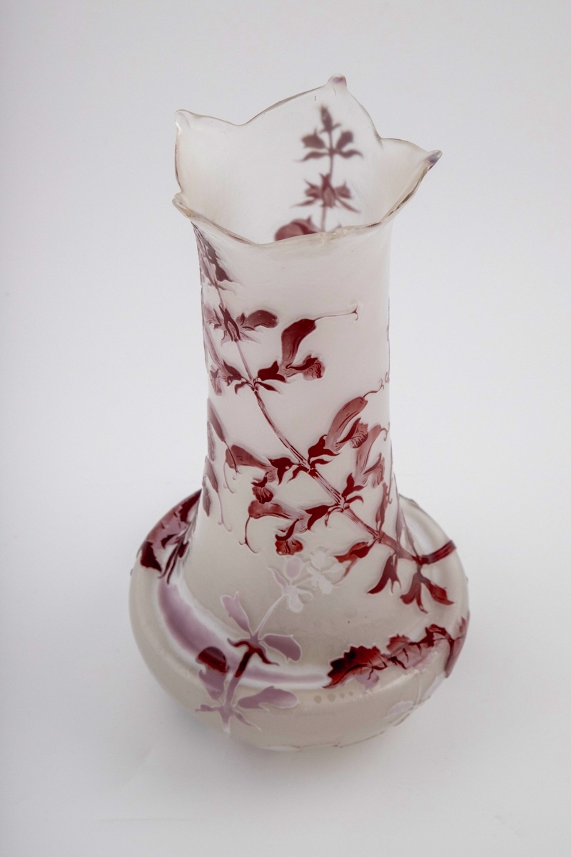 Høyreist vase i opakt melkehvitt glass. Nedre del av korpus er skålformet, hvorfra det reiser seg en høy sylinderformet hals. Vasens munning har en bladlignende utforming. Korpus er dekorert med overfangsteknikk, utformet som blomster og bladverk i lilla og burgunderrød. Dekoren er delvis bemalt med emaljemaling.