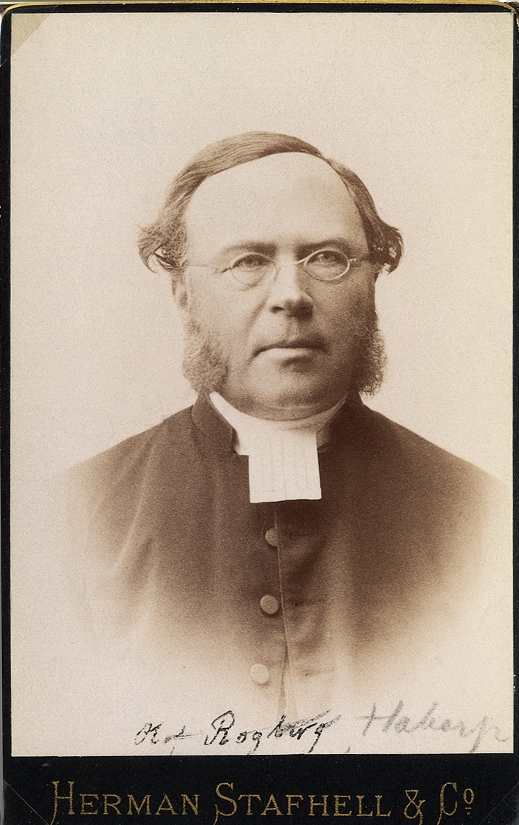 Foto av en man med glasögon och polisonger, klädd i prästrock med prästkrage. 
Knäbild, halvprofil. Ateljéfoto.