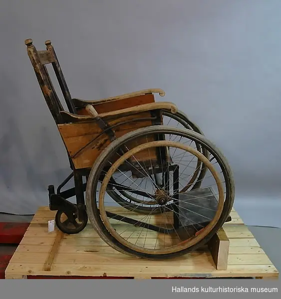 Rullstol av trä. Cykelhjul försedda med böjda trävidjor som används för att driva fram stolen. Fernissad. Däcken märkta: "Monark Extra". Hjul: diameter 60 cm."