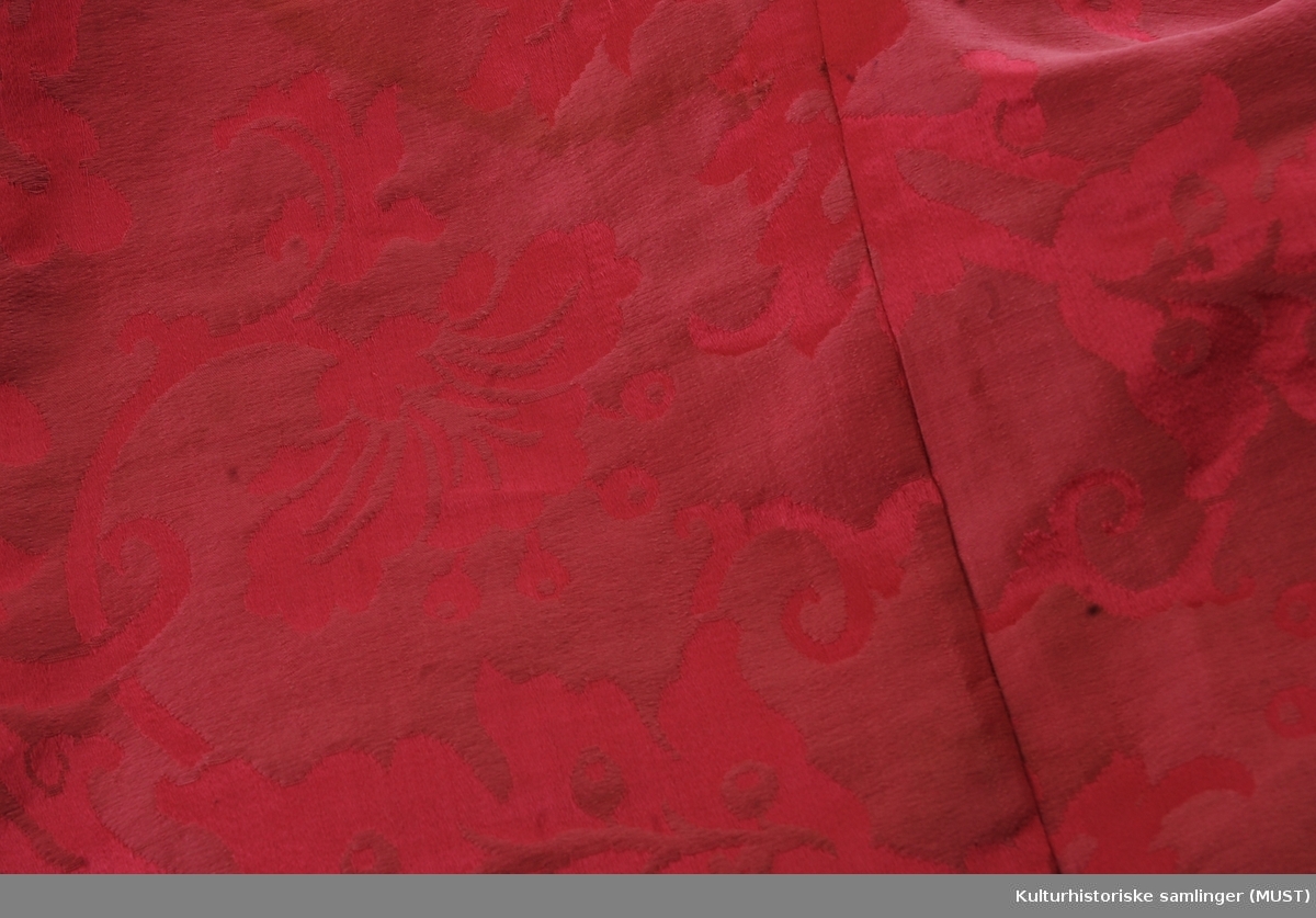 Kappe av rød silkedamask. Foret med sort bomullstøy med påtrykte blomster. Hette av samme stoff med blått for.
Snitt: To stykker med søm midt bak. Framme er det påsydd en forlenging som smalner av.
Stor hette sydd av fire stykker med midtsøm og buet søm fra 10cm framkant.
Dette stykket gir ekstra stoff til de mange foldene, elleve folder på hver side av midtsøm bak.
Kepen har av trykt bomullstoff (1700-talls)
Vattert med fin ullvatt. Innlagt hvit skrinnreim som tittekant mellom stoff og for rundt hele kapen. Samme hvite skinnkant påsydd rundt halsen.
