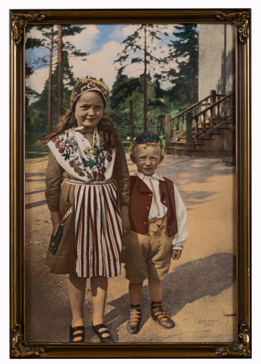 Fotografi, kolorerat, porträtt av två barn. Fotografiet är glasat och ramat med smal guldfärgad ram. Text på baksidan.