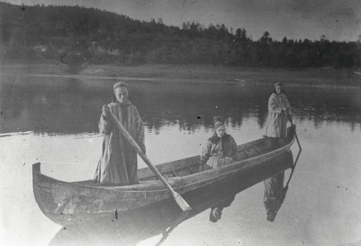 Motiv av tre kvinner i en kano/ elvebåt. Kvinnene har tradisjonelle drakter på seg (ládjogahpir og gákti). Kvinnen som sitter i båten er sannsynligvis Marie Øwre.