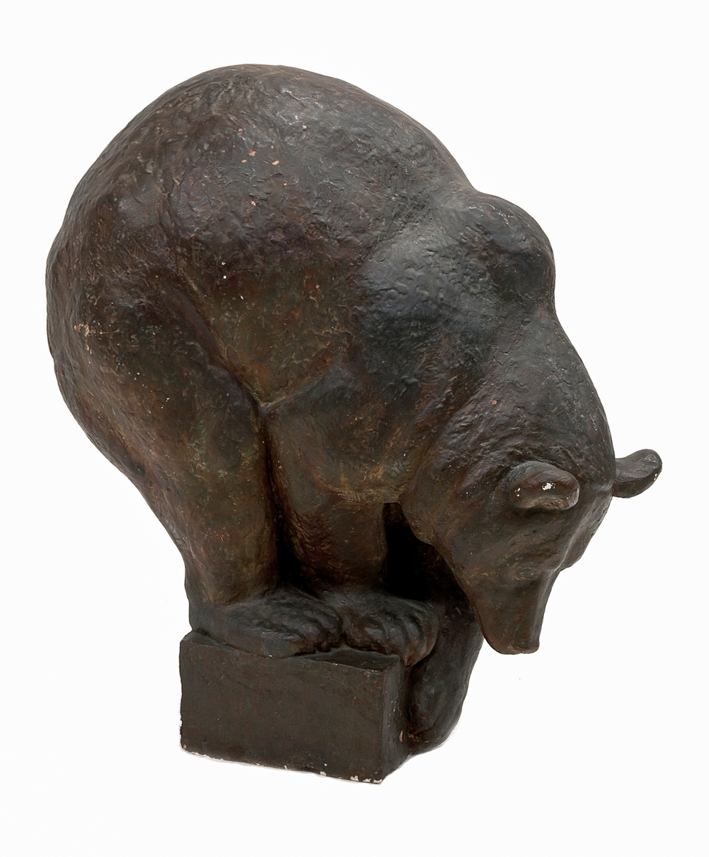 Skulptur, patinerad gips "Brunbjörn" av Arvid Knöppel. Björnen hopkrupen och framåtböjd med ena tassen på sockeln.