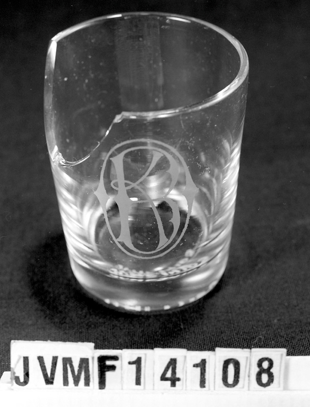 Dricksglas av klart glas med Ostkustbanans monogram ingraverat. Ett stort stycke av glaset saknas på ena sidan.