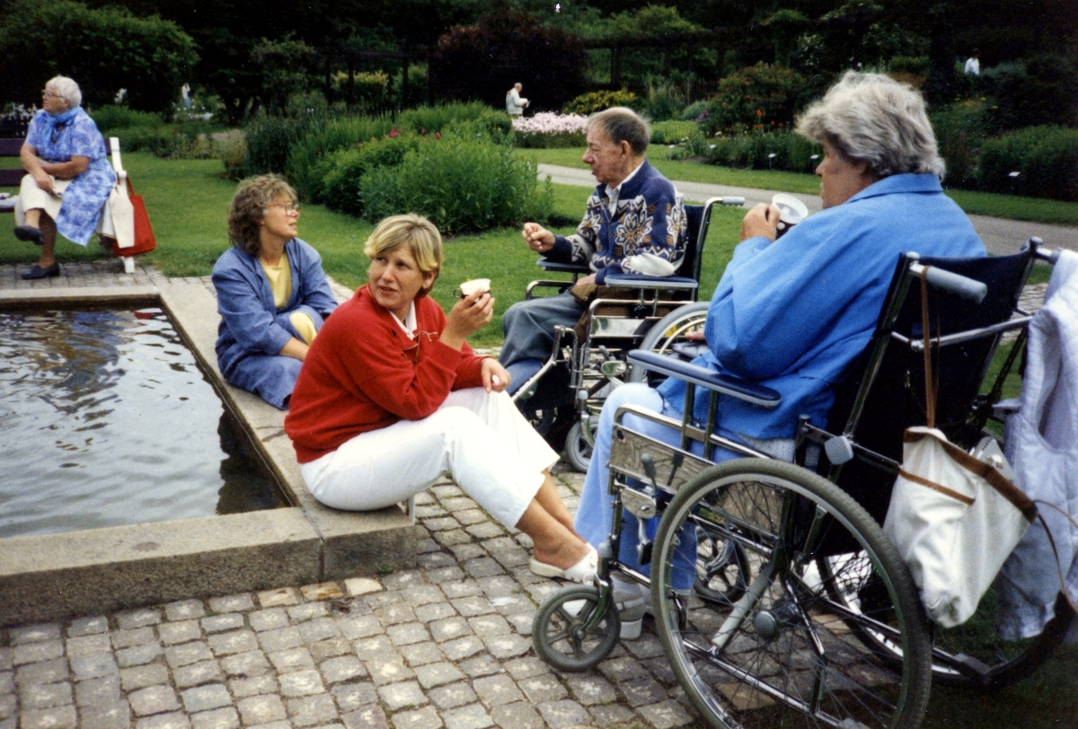 Brattåsgårdens äldreboende är på utflykt till Botaniska trädgården, 1980-tal. Från vänster: vårdbiträde Ingela Bengtsson (i röd tröja), vårdbiträde Anita Jansson (sitter på stenkanten), okänd man i rullstol samt Elsa Kristensson (i rullstol).