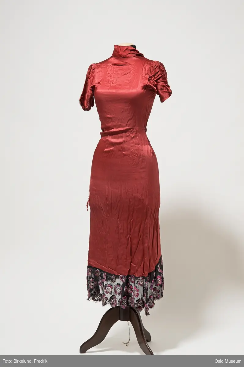 Mørk rød kjole i A-fasong. Korte ermer. Lukkes med gorv glidelås. Nede del av kjolen er kantet med et tynt sort stoff med blomsterdekor.
