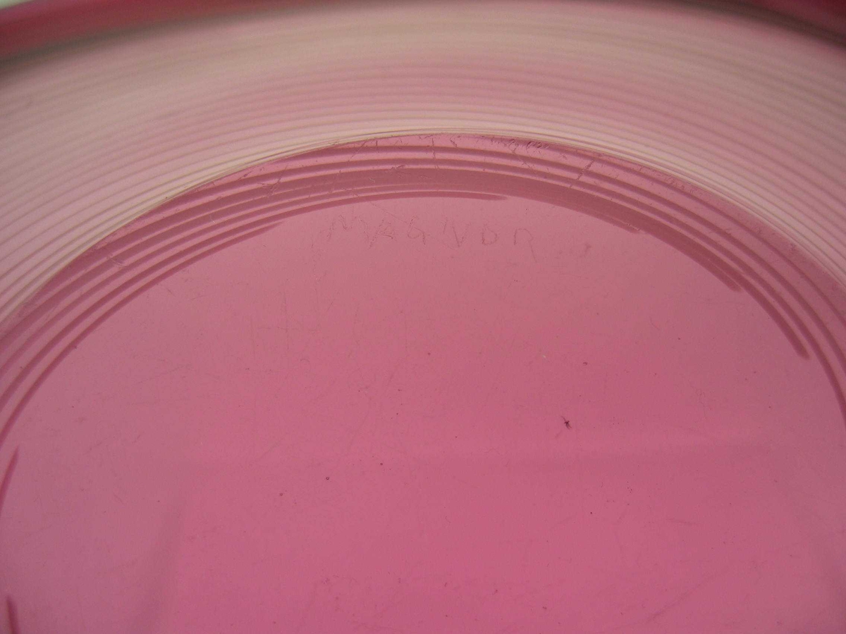 Avlangt fat på oval fotplate. Rødt glass med bølgete kant av klart glass. I det røde glasset en hvit spiral. 