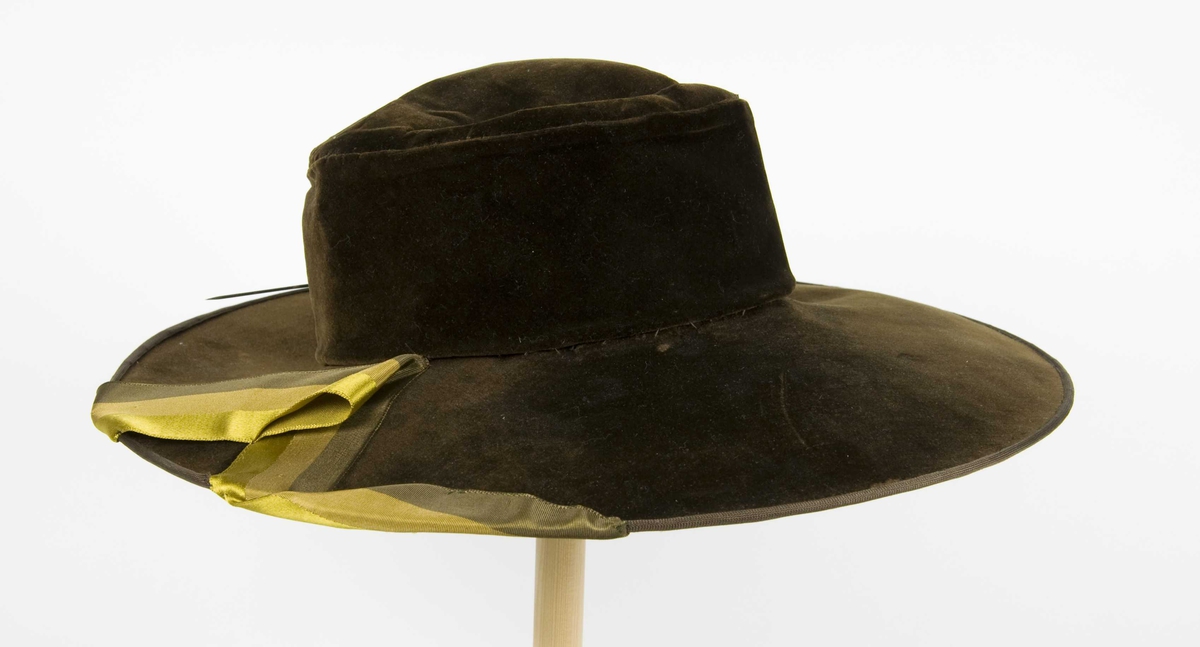Grønn plysjhatt med høy pull og bred brem. hatten har et trefarget påsydd silkebånd som pynt på den ene siden (virker sekundær).  Midt bak en hattebnål. Fôr av silke.