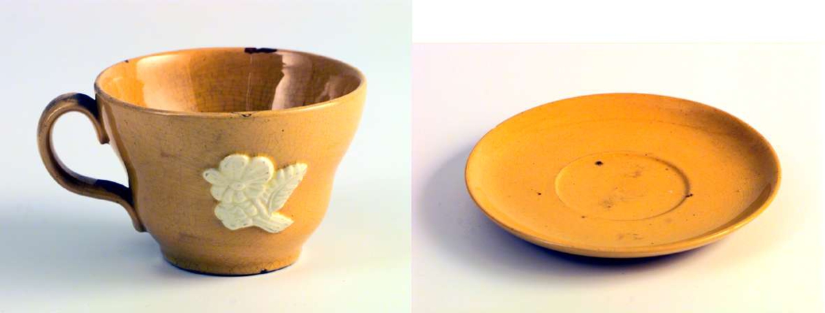 Gul kaffe-/tekopp med skål. Koppen er dekorert med en blomst med blad. Dekoren er hvit. Skålen er uten dekor. Det er hakk i både koppen og skålen.
Det er ingen produksjonsmerker på kopp eller skål.
