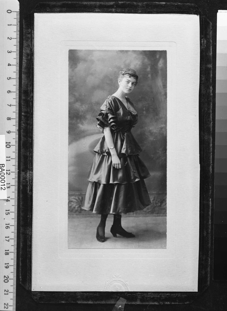 Atelierfotografi; helbilde av en ung dame i halvlang ballkjole med brede fleser i taft, sko med sløyfe og oppsatt frisyre. Bakgrunnsteppe viser en parksti kantet med trer og busker i solskinn.