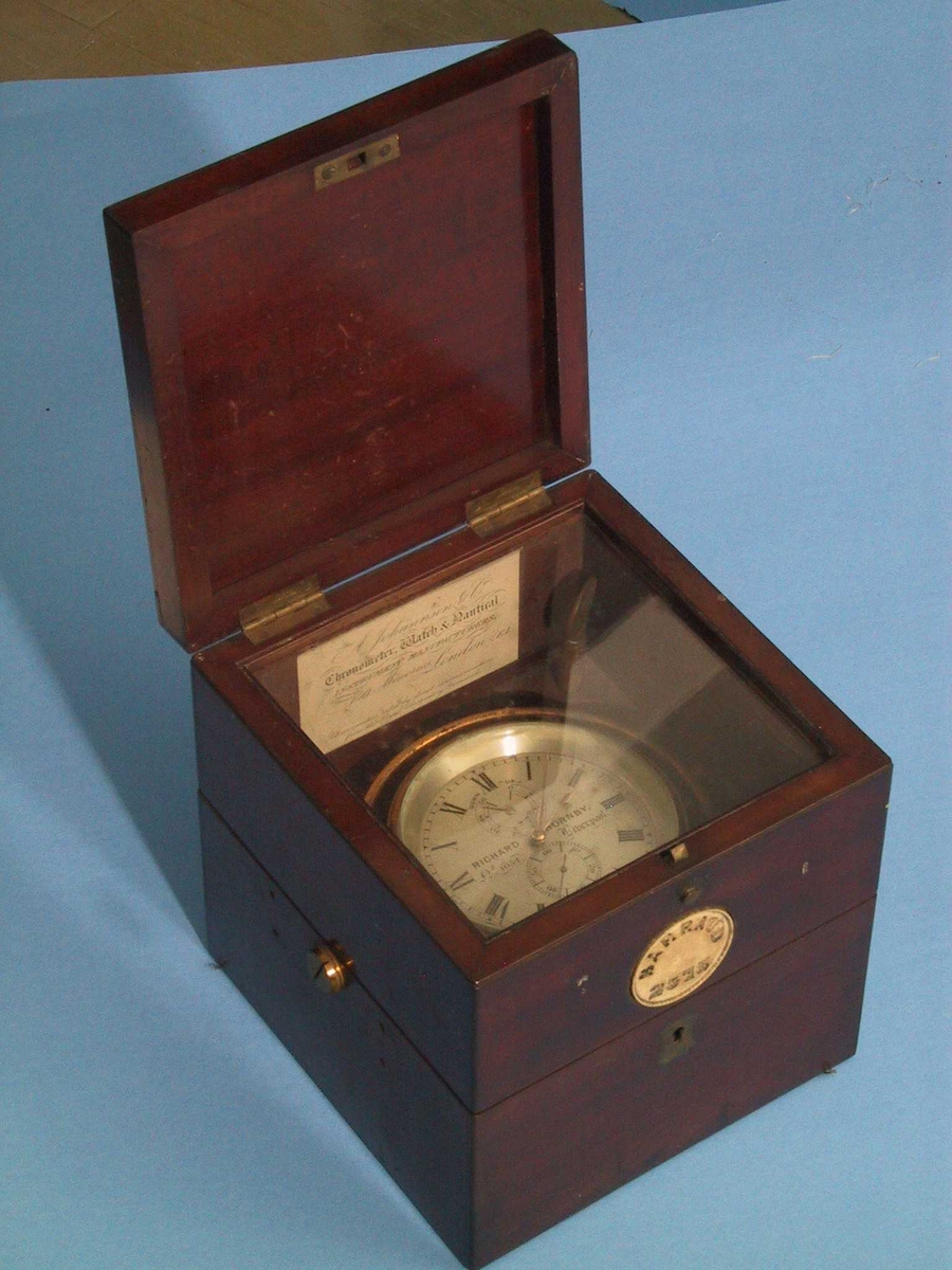 a) Kronometer i kasse av   mahogni  med messing  beslag.   Metall  med messingbelegg,   glass.   a)H.18.Bunn 17,1x17,5. Diam.kron.10,7. b)H.24,4.Bunn L.31,2.  B.24,4.  b) Ytre kasse,   palisander  med stopning innv.av puter trukket med seilduk og seilduksgarn. a) Firkantet kasse med to lokk, det øverste er smalt, under dette en  glasskive gjennom hvilken en kan se kronometret. Det annet lokk deler  etuiet i to. Kronometer i metallring, alt med messingbelegg, som  dels flasser av, firmanavn på skiven har romertall:   Tilst. Febr. 1967: metallet litt anløpet, ellers god  b) Firkantet kasse med lokk som deler kasse i to, messingkrok. På  sidene et lite buet felt i bunnen med hull for skrue. Midt i sidene  festet en rem, som festet over lokket. Innvendig puter i bunn og  side. Flg. papirer lå i bunnen:  1) Kvittering for kjøpet, datert August 8th 1923. har ikke pris =  garantiseddel.  2) Firmakort fra "Alf. Willings & Co. Marine Opticians and Compass  Adjusters.73, Church Street, West Hartlepool . Authorized Compass  Adjusters for Norwegian Vessels at this Port by Appointment of the  Norwegian Government."  3) En pappskive med blå blyant. ORA.  4) Papir med avlest kronometer for juni, juli og aug. 1935. øverst:  Kronometer no. 1054 "Ora".  5) Ditto for 9. febr. 1926. o.a. datoer i febr.  6) Ditto for 15/9.  