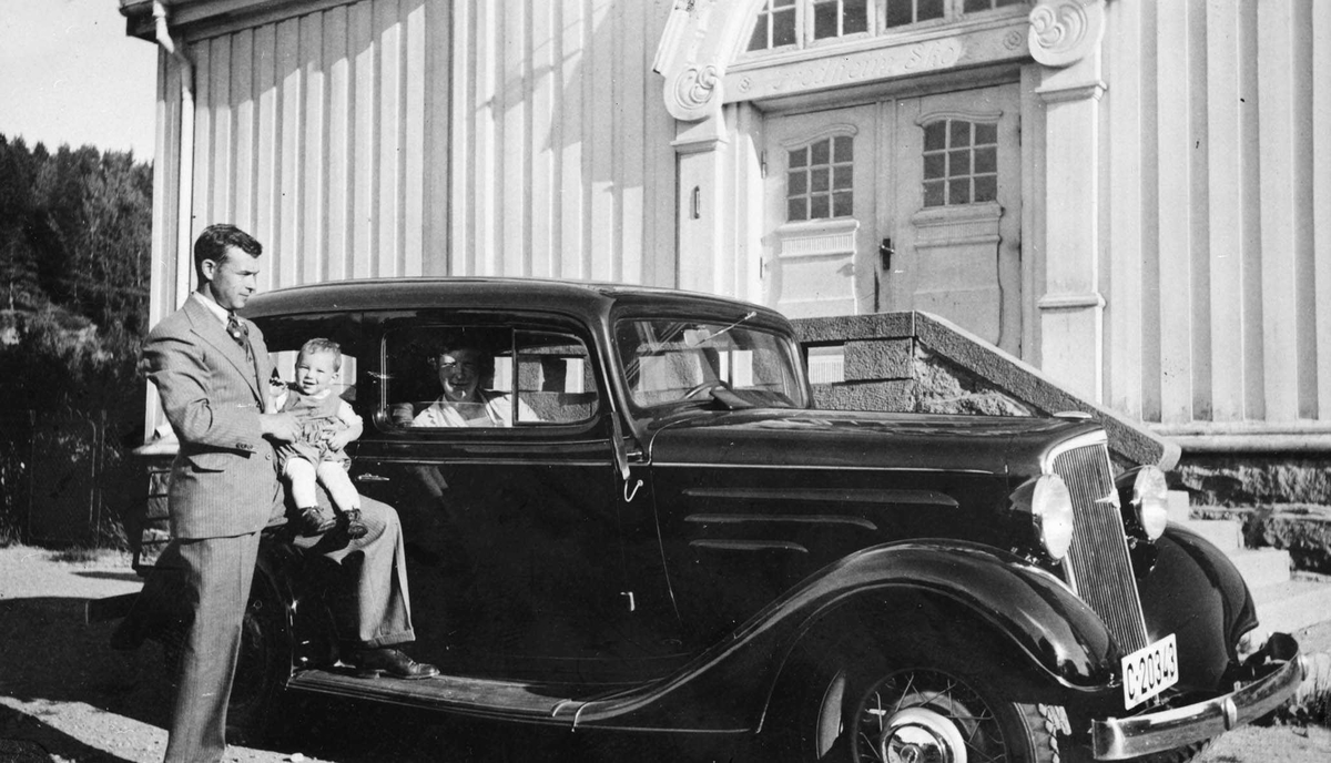 Bil foran inngangen på Fredheim skole. Mann i dress med liten gutt på kneet, kvinne passasjer i bilen.