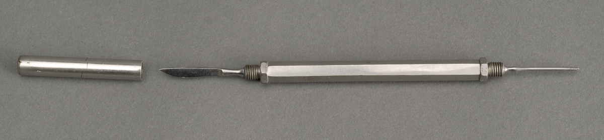 6 kantet metallholder med corneaskrape og liten kniv, skalpell, i hver sin ende. Beskyttelselokk med skru i den  ene enden.